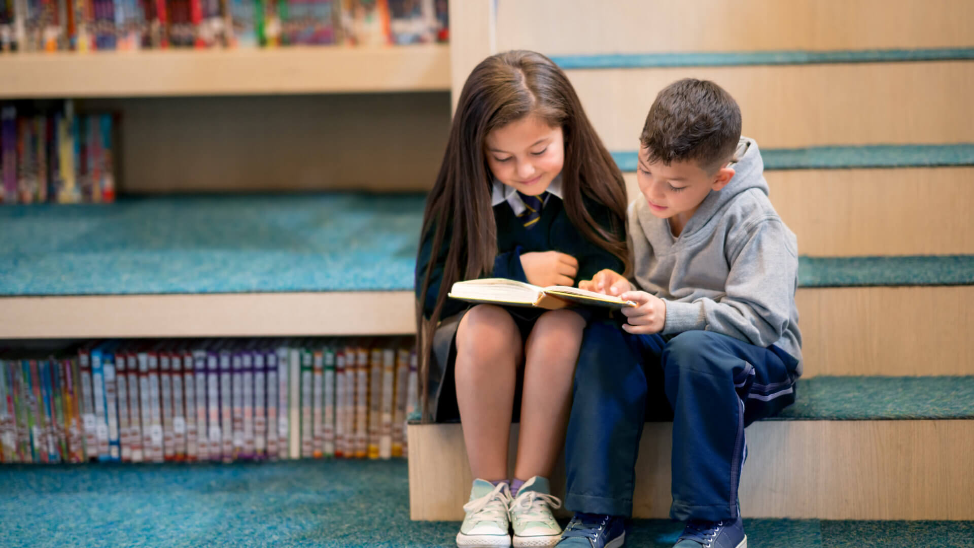 2 young students reading a book together on the library stairs.