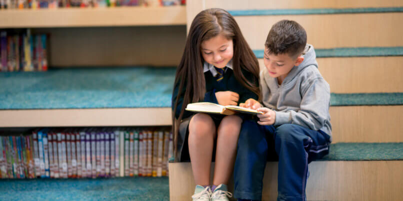 2 young students reading a book together on the library stairs.